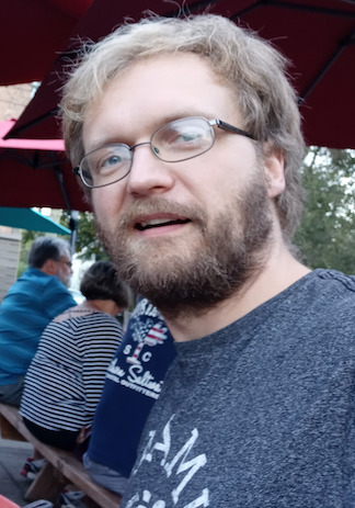Hunter, wearing a slate grey shirt. The shirt has partial text visible, reading Myaamia, and he is facing toward the camera and smiling with mouth open, at a restaurant's outdoor patio.