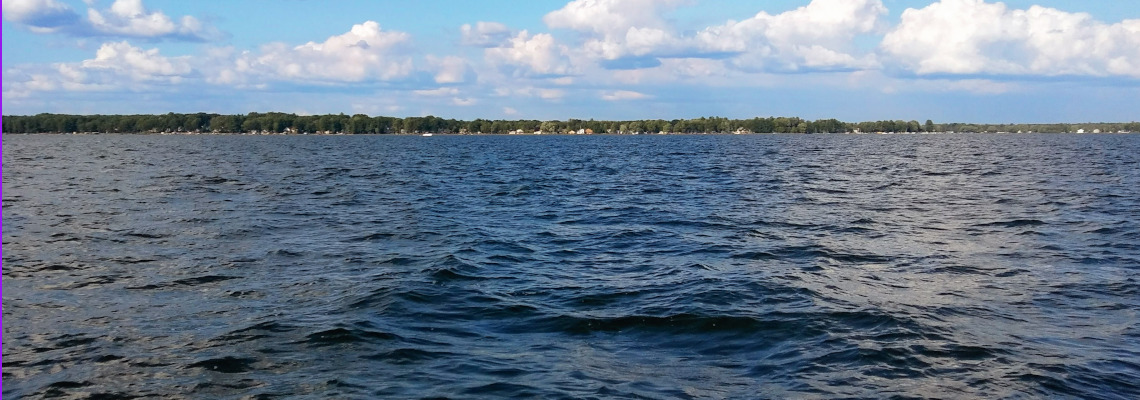 Puffy clouds populate a blue sky over the shores of Houghton Lake, Michigan, as viewed from out in the water